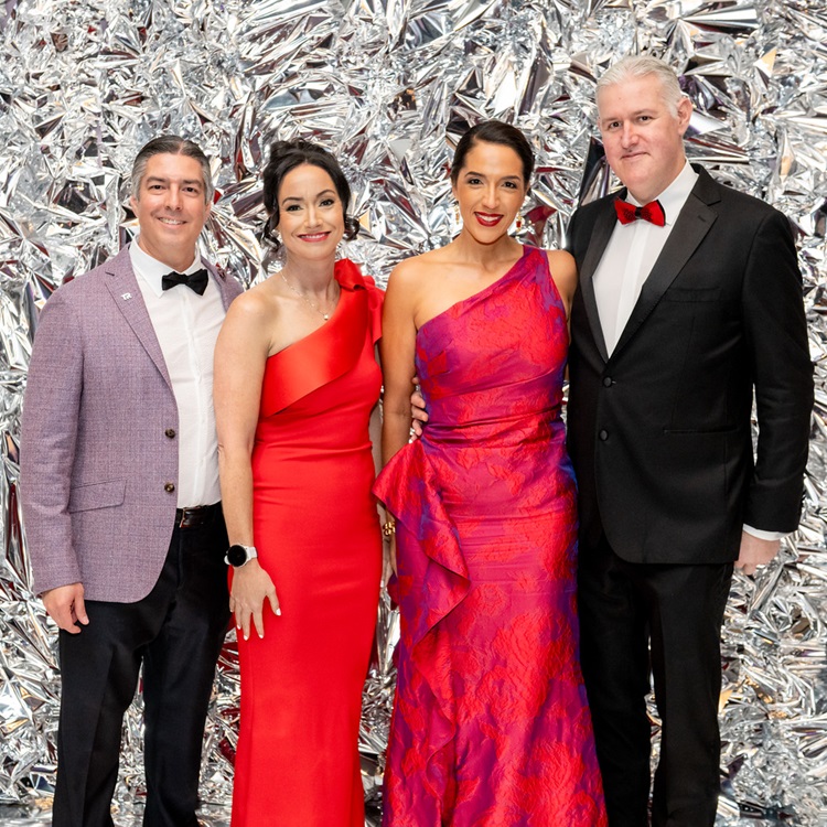 Two couples in formal wear standing in front of a silver metallic backdrop.